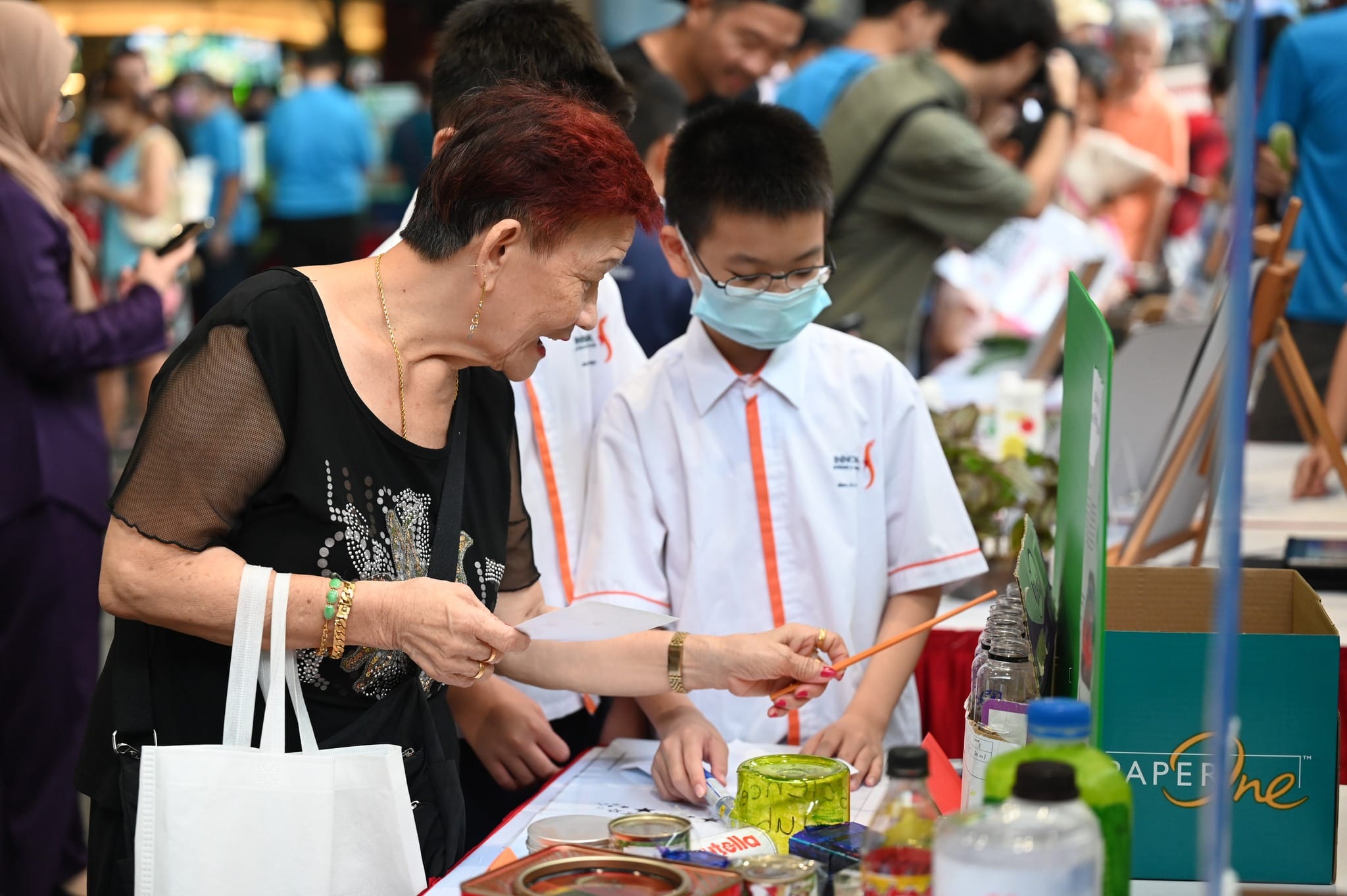 An older woman with red hair holds a paintbrush at a craft table while a masked schoolboy in uniform watches nearby.